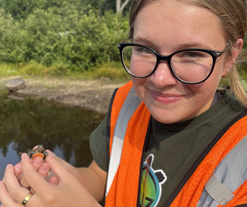 A young white woman in a Turtle Guardians t-shirt and orange reflective vest beside a small body of water holding a turtle hatchling in her hand.